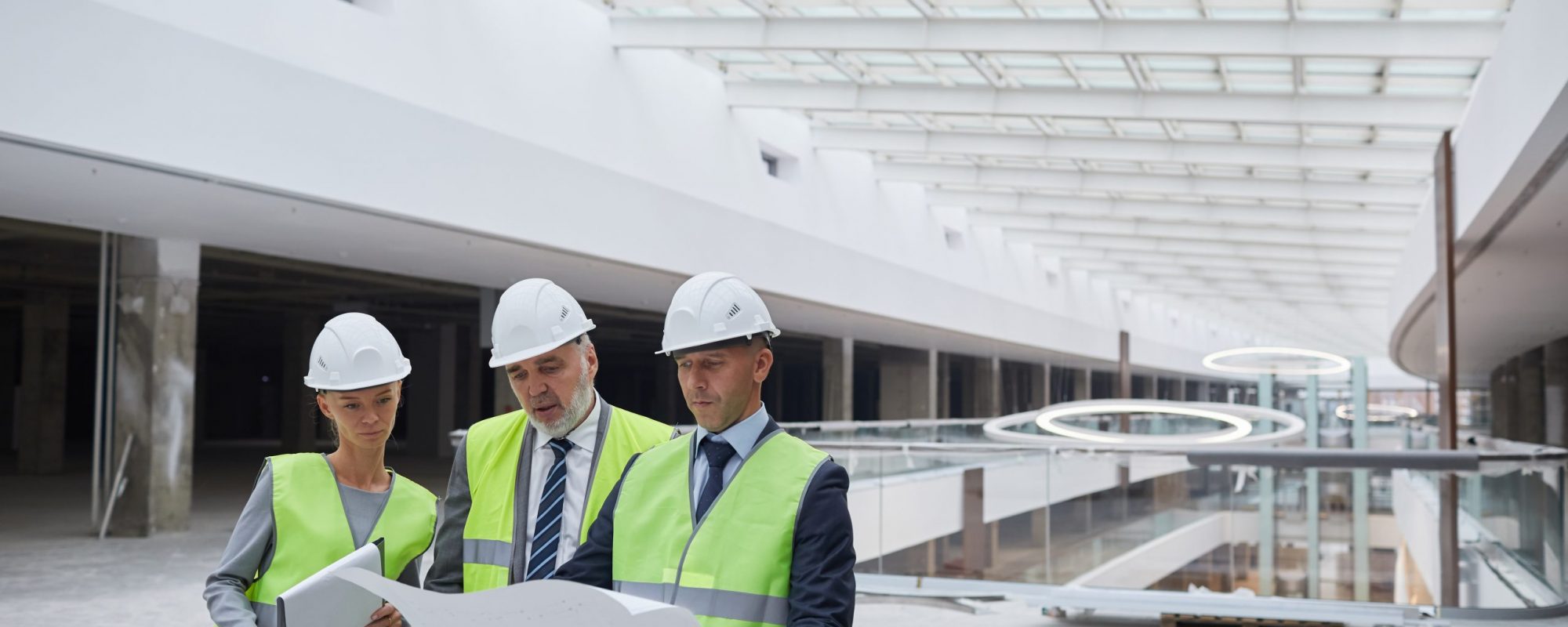 Waist up portrait of three successful business people wearing hardhats and inspecting blueprints while standing at construction site indoors, copy space